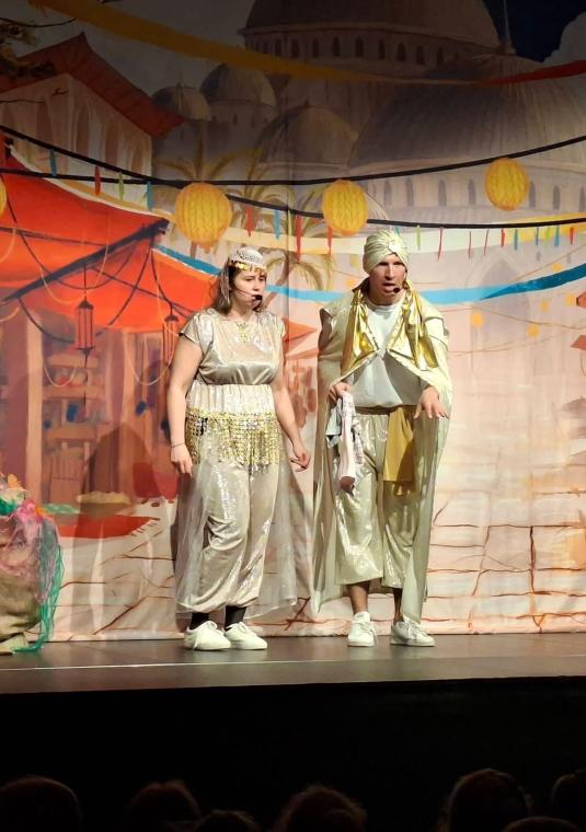 Kadr ze spektaklu spektaklu teatralnego przedstawiający dwoje aktorów w orientalnych strojach na scenie. Z lewej strony stoi kobieta w jasnej sukni z ozdobnym pasem, z prawej mężczyzna w turbanie i pelerynie. W tle widać malowaną scenografię przedstawiającą orientalny targ pod kopułami pałaców oraz wiszące lampiony i girlandy.