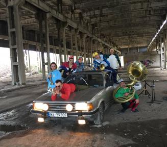 Grupa muzyków z instrumentami dętymi, pośrodku samochód osobowy, dwóch muzyków leży na samochodzie, nad nimi konstrukcja mostu.