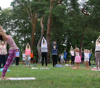 Grupa kilkunastu kobiet w pozycjach z wyciągniętymi rękoma podczas ćwiczenia jogi, w parku wśród drzew, kobiety w strojach sportowych, obok rozłożonaematy do ćwiczeń