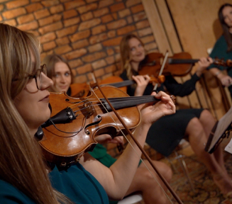 Cztery młode kobiety. Siedzą na krześle. Każda trzyma instrument smyczkowym, na którym gra.