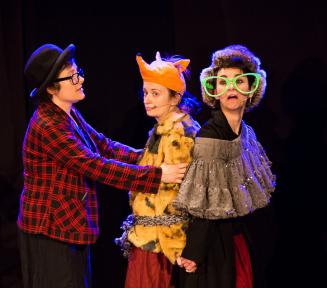 Trzy młode kobiety - aktorki stoją obok siebie na scenie. Jedna kobieta ubrana jest w marynarkę w czarno-czerwoną kratę, na głowie ma czarny kapelusz, na nosie czarne okulary. Druga kobieta ma na głowie czapkę z głową pluszowego lisa, na sobie żółte futro. Trzecia kobieta na nosie ma duże zielone okulary, na głowie zimową czapkę. W tle czarne tło.