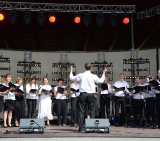 Kilkanaście śpiewających osób stoi na scenie, w półkolu, każdy odświętnie ubrany, każdy trzyma przed sobą otwarty zeszyt z nutami, przed nimi młody mężczyzna z podniesionymi rękami, dyrygujący zespołem, nad nimi widoczne oświetlenie sceniczne