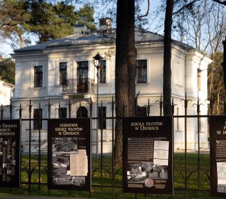 Na pierwszym planie płot z kutej stali w kolorze czarnym, na nim zawieszone plansze z napisami i zdjęciami.