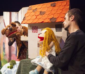 Bajkowa scenografia teatralna, na scenie aktor który w rękach trzyma duże lalki szmaciane. 