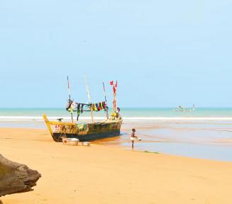 Zdjęcie plaży nad brzegiem morza. Przycumowana jest przy niej łódź rybacka, obok stoi chłopiec w kapielówkach.