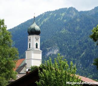 Biała wieża kościoła na czubkami zielonych drzew. Za nia zalesiona góra. 