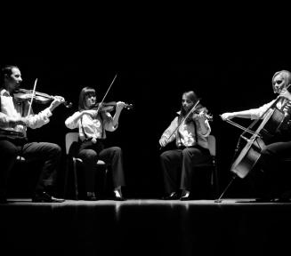 Biało-czarne zdjęcie czterech osób ze skrzypcami w rękach. Jedna z nich ma wiolączelę. są to trzy kobiety i jeden mężczyzna. Siedzą na krzesłach w półkolu.