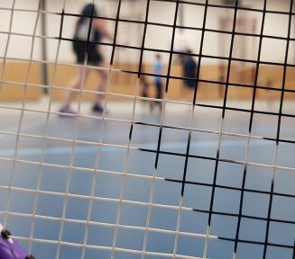 Na zbliżeniu siatka rakietki do badmintona. Przez nią widać sylwetki trzech osób na sali gimnastycznej.