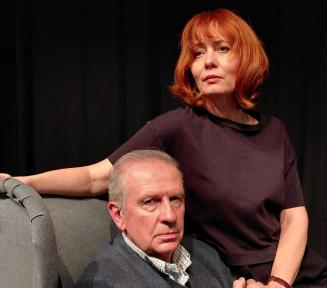 Zdjęcie kobiety i mężczyzny siedzących na szarym fotelu. Czarne tło.