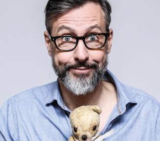 zdjęcie portretowe Szymona Majewskiego, patrzy na nas z figlarnym uśmieszkiem, w rękach trzyma starego, białego pluszowego misia