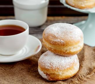 zdjęcie apetycznych pączków obsypanych cukrem pudrem i porcelanowa filiżanka z herbatą, w tle w nieostrości, platera z pączkami i cukiernica