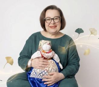 Uśmiechnięta kobieta w okularach i ciemnozielonej bluzce siedzi, trzymając na kolanach tradycyjną, słowiańską motankę. Lalka ubrana jest w białą koszulę, czerwony gorset, wzorzystą spódnicę z koronkowym fartuszkiem oraz chustę na głowie. Wokół postaci widoczne są graficzne elementy w postaci złotych i zielonych liści miłorzębu, które zdają się unosić na świetlistych liniach.