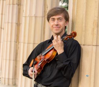 Mężczyzna o jasnej karnacji w czarnej koszuli, trzymający skrzypce i smyczek, opierający się o klasyczną kolumnę.