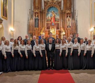 Konstanciński Chór Kameralny na wspólnym zdjęciu wewnątrz kościoła. Kilkudziesięcioosobowy zespół ustawiony jest w dwóch rzędach przed ozdobnym, drewnianym ołtarzem. Kobiety ubrane są w białe koszule i długie czarne spódnice, a mężczyźni w czarne garnitury z muchami.