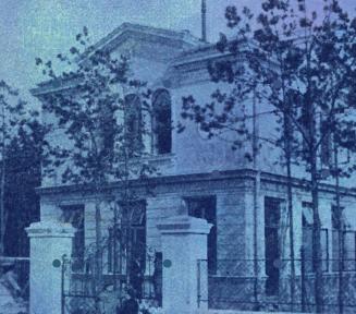 Archiwalna fotografia zabytkowej, piętrowej willi w Konstancinie, utrzymana w monochromatycznej, niebieskiej tonacji. Budynek o klasycznej architekturze z ozdobnymi gzymsami jest otoczony drzewami i ozdobnym ogrodzeniem z murowanymi słupkami na pierwszym planie.
