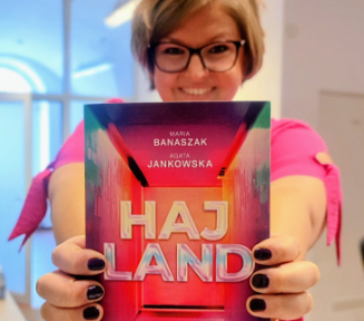 Zdjęcie uśmiechniętej kobiety, która trzyma przed sobą książkę, na okładce napis Haj Land