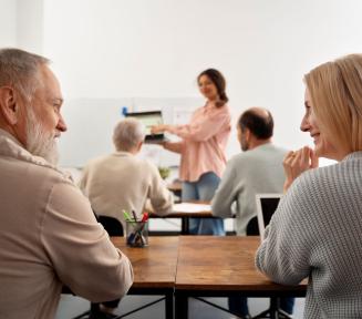dwoje ludzi siedzi przy stole i patrzą na siebie, w tle widać nauczycielkę trzymającą laptopa