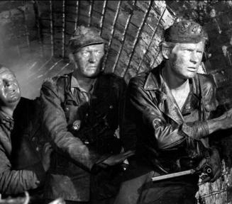 Czarno-biały kadr z filmu „Kanał” Andrzeja Wajdy przedstawiający trzech brudnych, wyczerpanych powstańców w wojskowych furażerkach, skradających się w wąskim, ceglanym kanale ściekowym.