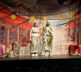 Kadr ze spektaklu spektaklu teatralnego przedstawiający dwoje aktorów w orientalnych strojach na scenie. Z lewej strony stoi kobieta w jasnej sukni z ozdobnym pasem, z prawej mężczyzna w turbanie i pelerynie. W tle widać malowaną scenografię przedstawiającą orientalny targ pod kopułami pałaców oraz wiszące lampiony i girlandy.
