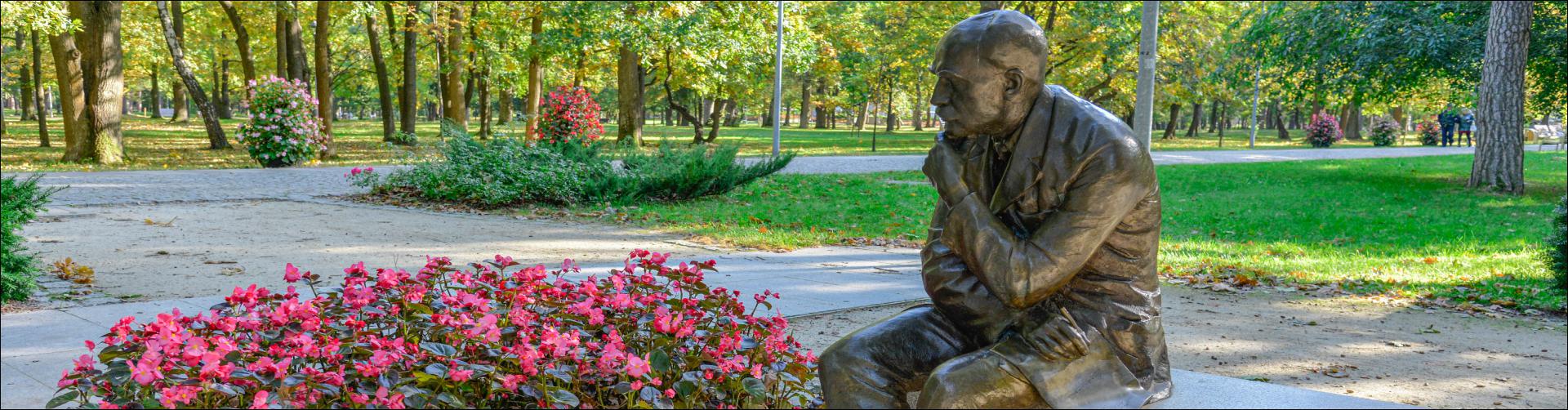 Park Zdrojowy. Pomnik Stefana Żeromskiego przedstawiający pisarza siedzącego na jednej z ławek. Obok rosną różowe kwiaty. W tle widać aleje spacerową, obok której rosną zielone drzewa. 